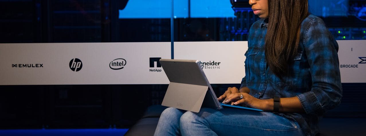 Mulher com óculos e camisa xadrez sentada, trabalhando em um tablet com teclado em um ambiente de data center com luzes azuis e logotipos de empresas de tecnologia ao fundo.