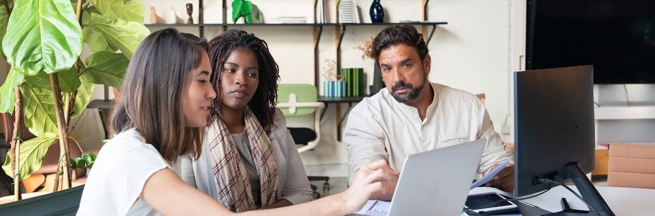 Três profissionais em uma sala de reunião organizada. Uma mulher à esquerda aponta para a tela de um notebook que mostra gráficos e códigos, enquanto uma mulher e um homem com barba observam atentamente. Ao fundo, uma estante com objetos decorativos e plantas.